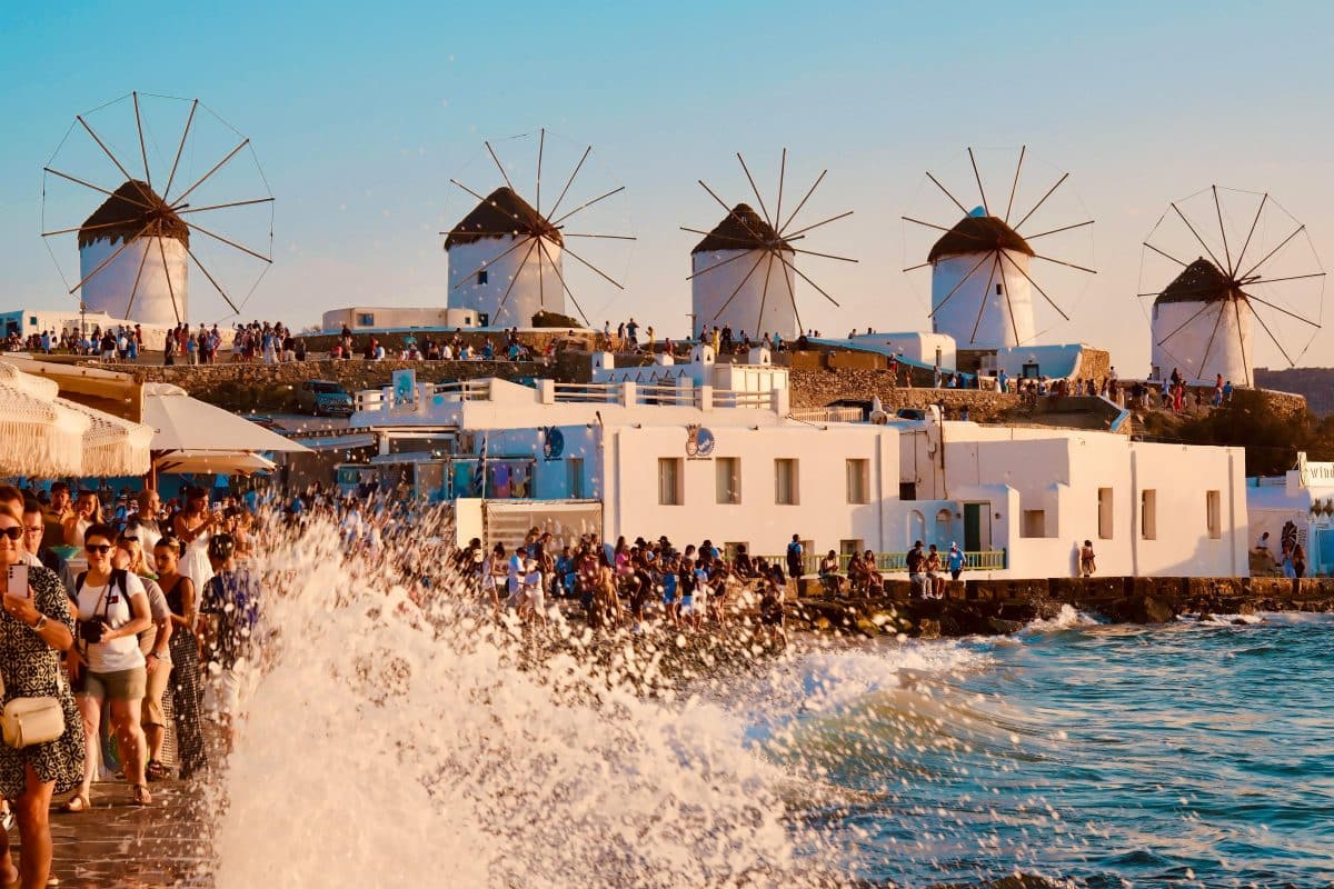 Tourists walking along the waterfront near the famous windmills of Mykonos as waves crash against the promenade at sunset.