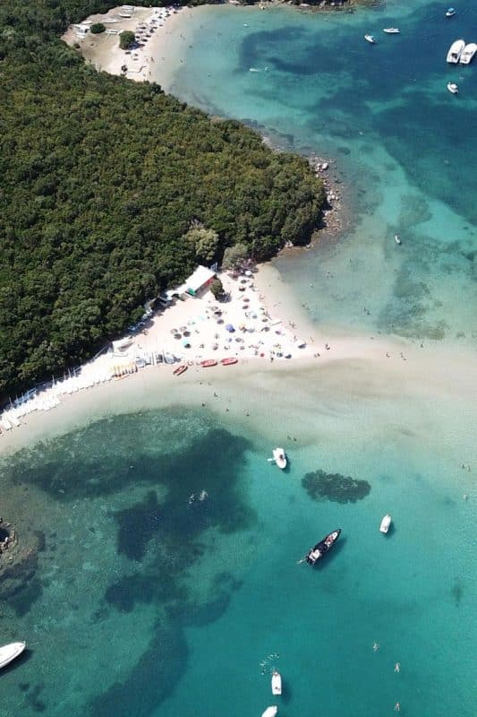 Aerial view of Bella Vraka Beach in Syvota, Greece, showing the shallow sandbar connecting the mainland to Mourtemeno Island.
