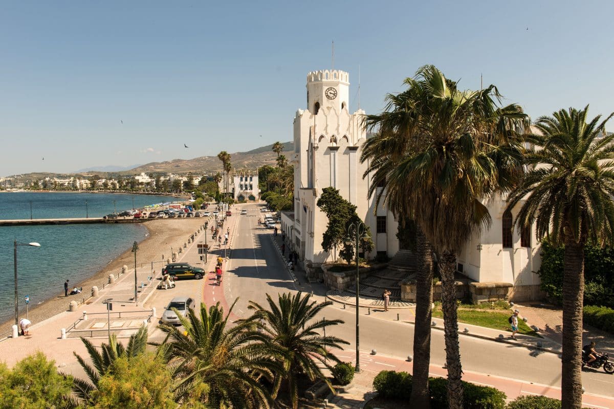 View of Kos Town's waterfront lined with palm trees, a historic building, and the Aegean Sea along the promenade.