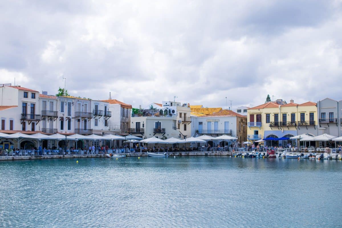 View of Rethymno's waterfront harbor in Crete with colorful historic buildings, waterfront restaurants, and calm sea waters.
