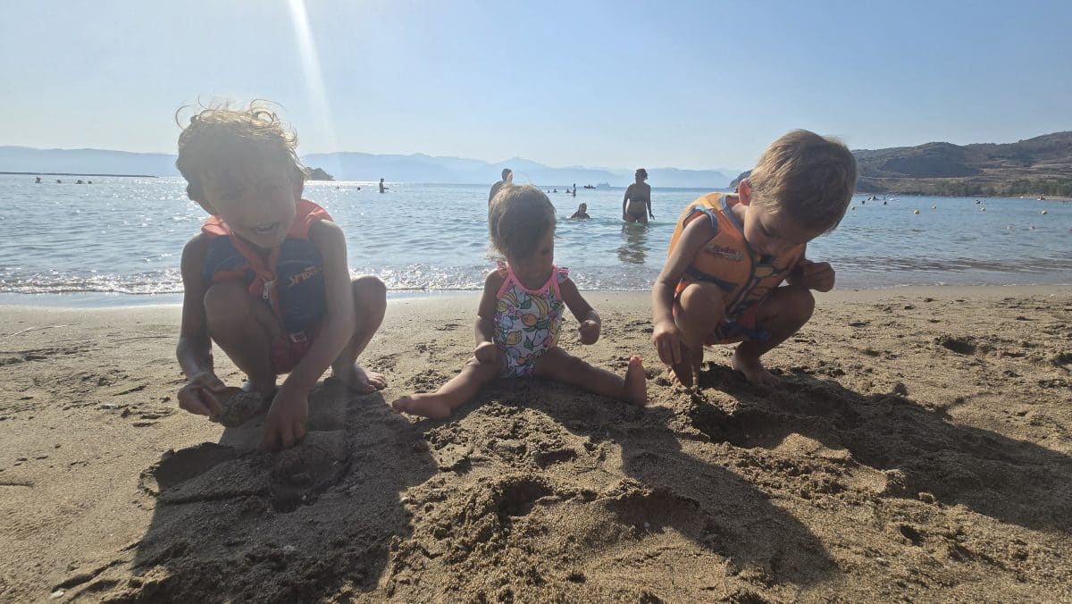 Three young children playing in the sand at Karathona Beach near Nafplio, Greece, a calm and family-friendly beach with shallow water.