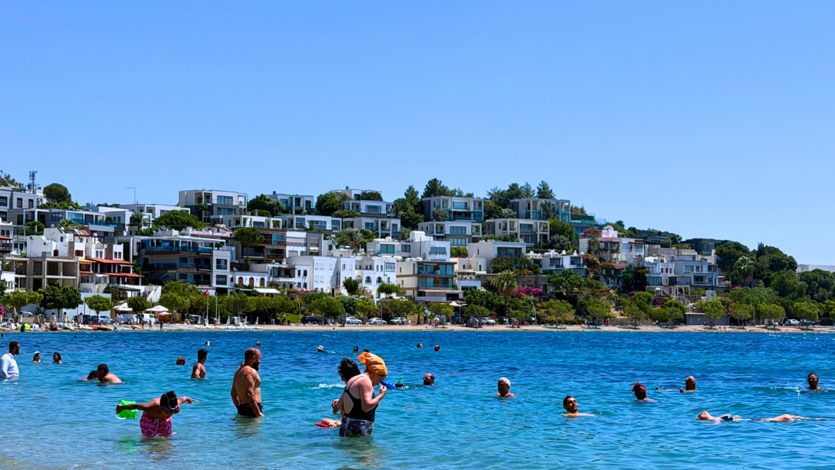 People swimming in the clear blue water at Kumbahçe Beach in Bodrum, Turkey, with white buildings along the waterfront.